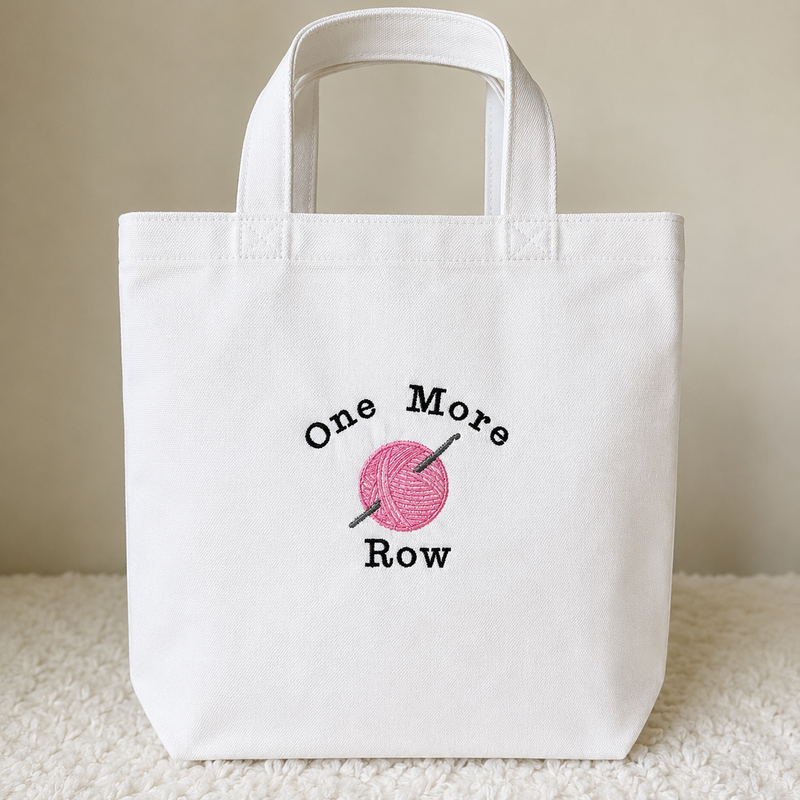 A white canvas tote bag with a pink and black embroidered design reading 'One More Row' around a pink ball of yarn with a knitting needle, set against a neutral beige background.