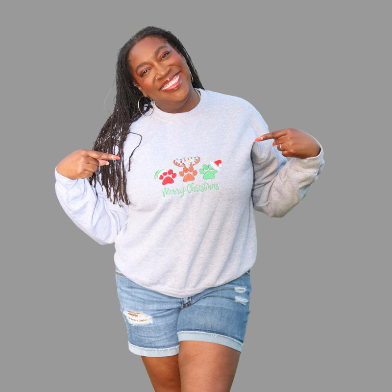 A woman standing wearing a light gray heather crewneck sweatshirt with a colorful Christmas paw-print graphic and the words 'Merry Gathering' on the front, paired with distressed denim shorts.