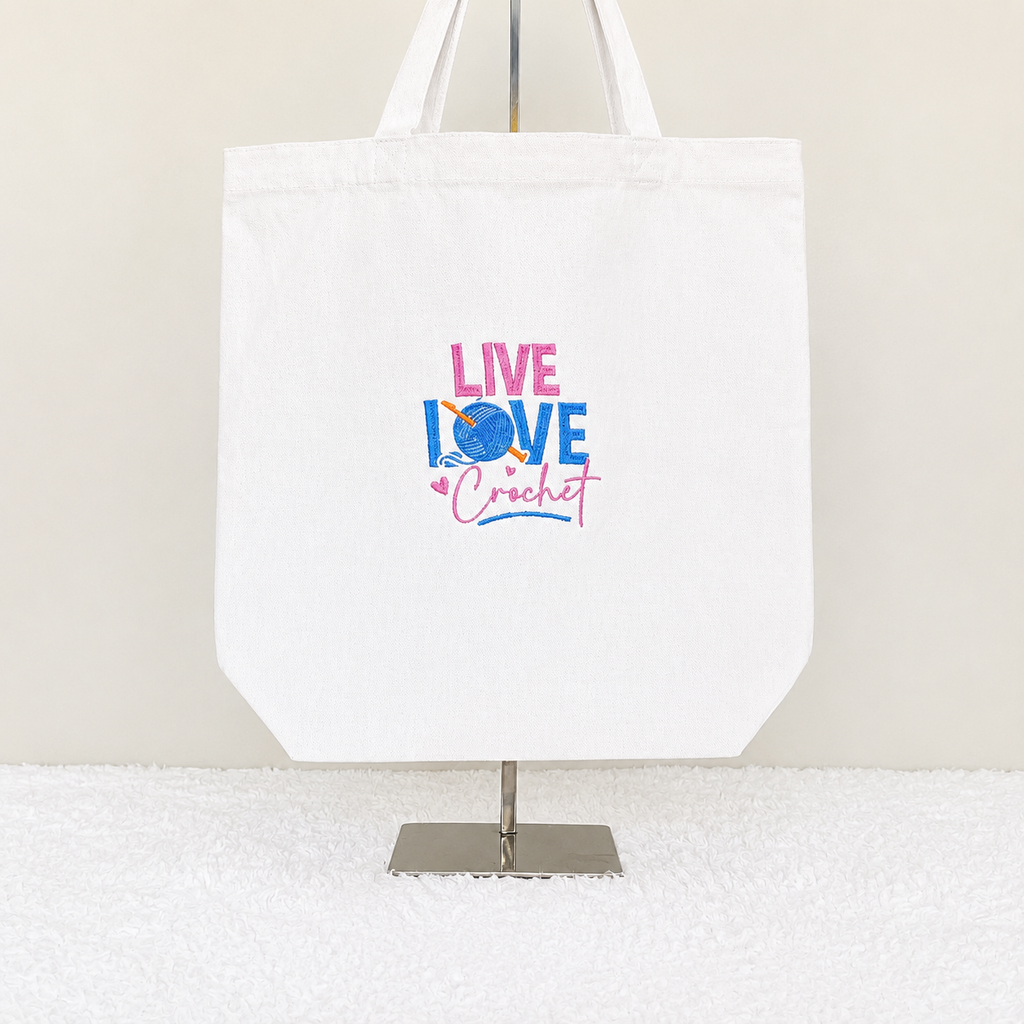 A white canvas tote bag displayed on a metal stand against a light beige wall and white textured surface. The bag features a colorful graphic in the center that reads 'LIVE LOVE Crochet' in pink and blue lettering with a crochet hook and ball motif.