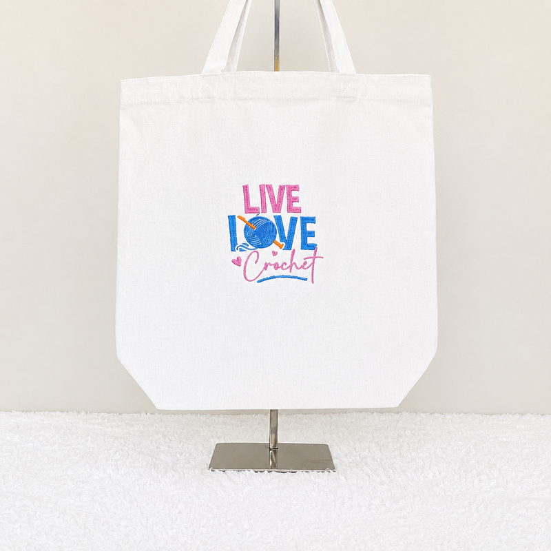 A white canvas tote bag displayed on a metal stand against a light beige wall and white textured surface. The bag features a colorful graphic in the center that reads 'LIVE LOVE Crochet' in pink and blue lettering with a crochet hook and ball motif.
