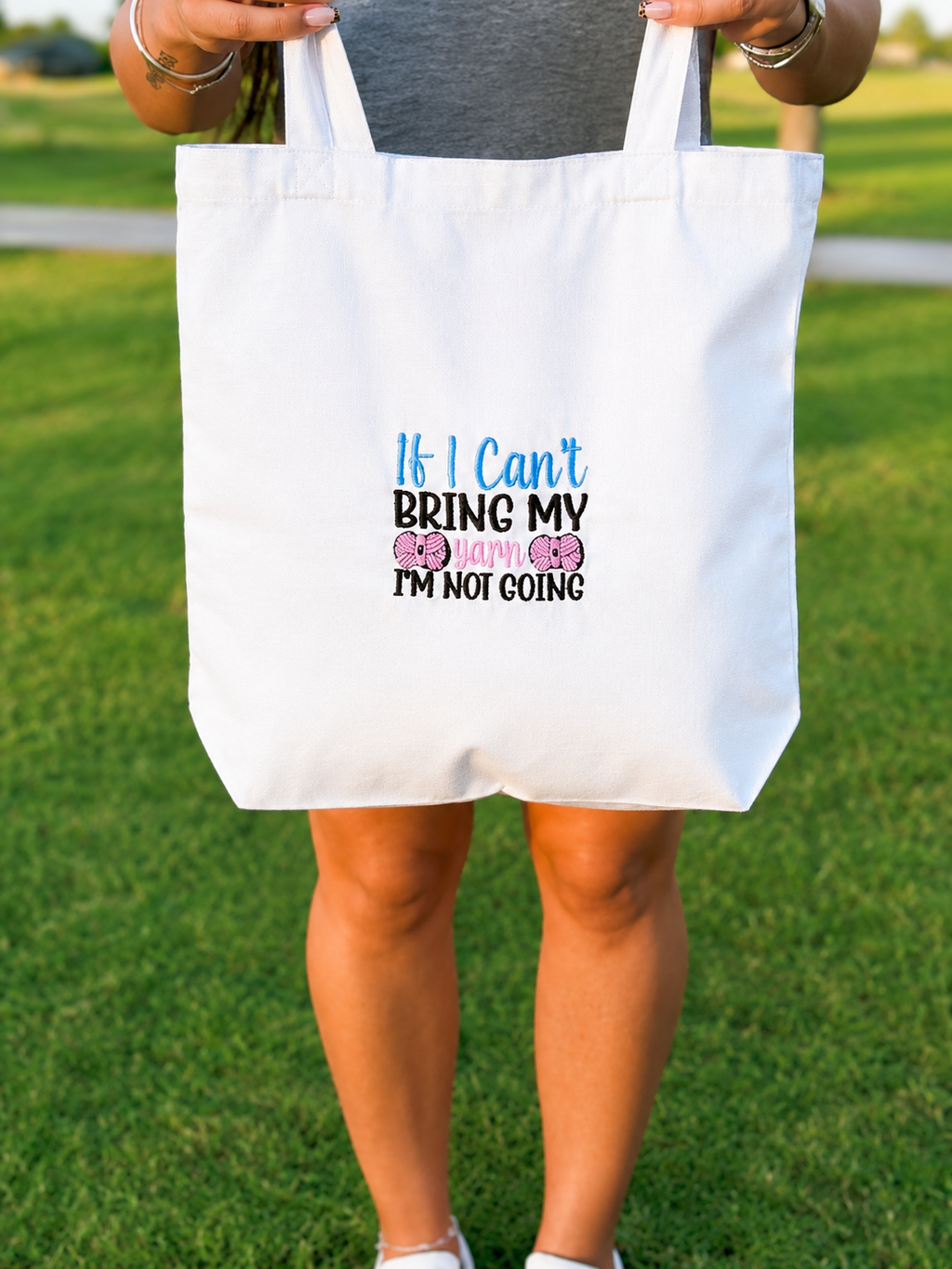 A white canvas tote bag held by a person outdoors on a grassy lawn. The bag features a printed message that reads 'If I Can't BRING MY mom and dad I'm NOT going' with pink and blue heart graphics.