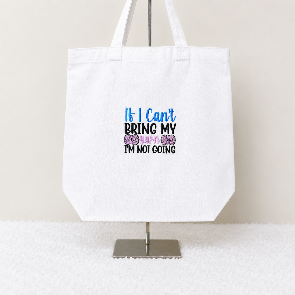 A white canvas tote bag displayed on a metal stand against a light beige wall and white textured surface. The bag features a printed slogan that reads 'If I Can't BRING MY BABY I'M NOT GOING' in blue, black, and purple text with two purple bows.