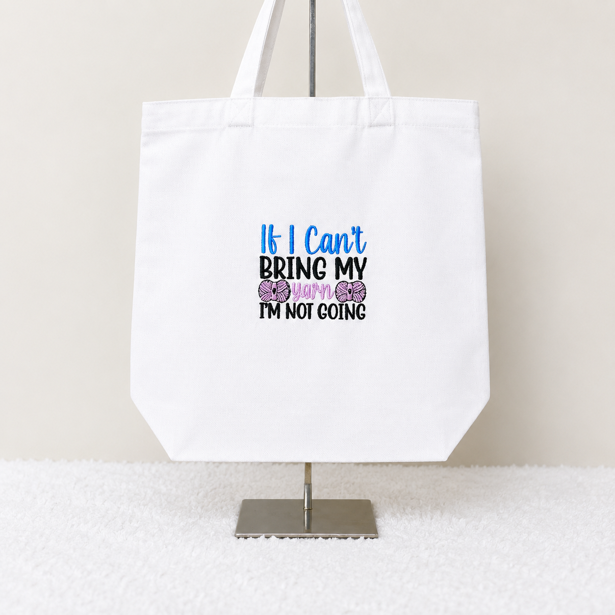 A white canvas tote bag displayed on a metal stand against a light beige wall and white textured surface. The bag features a printed slogan that reads 'If I Can't BRING MY BABY I'M NOT GOING' in blue, black, and purple text with two purple bows.