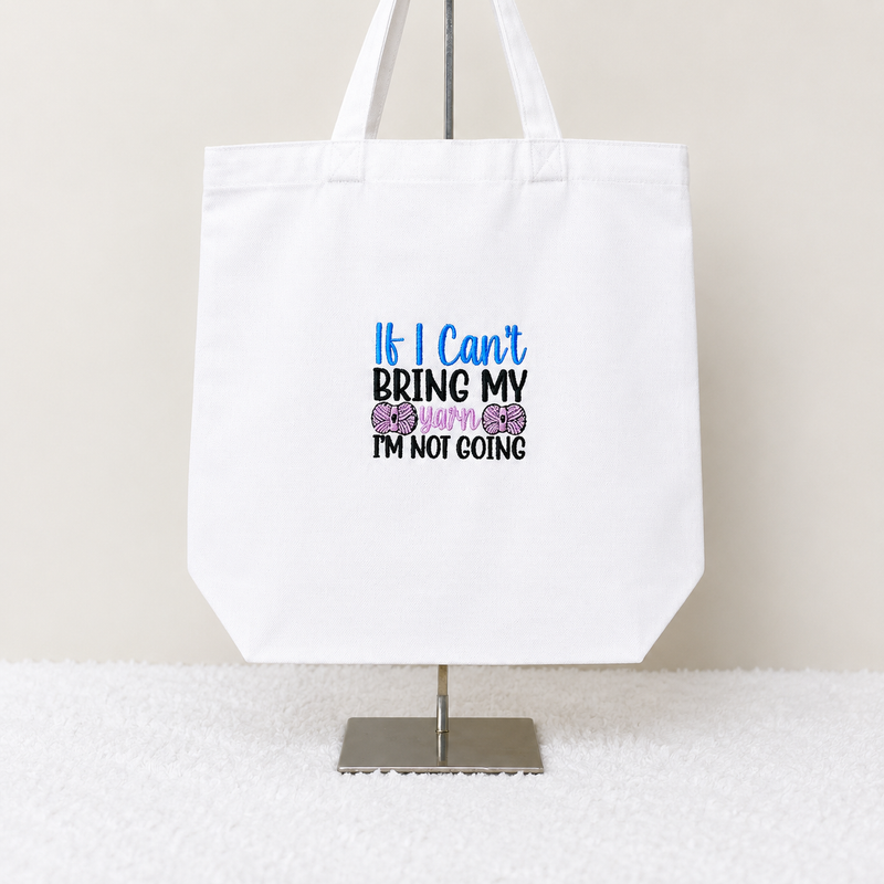 A white canvas tote bag displayed on a metal stand against a light beige wall and white textured surface. The bag features a printed slogan that reads 'If I Can't BRING MY BABY I'M NOT GOING' in blue, black, and purple text with two purple bows.