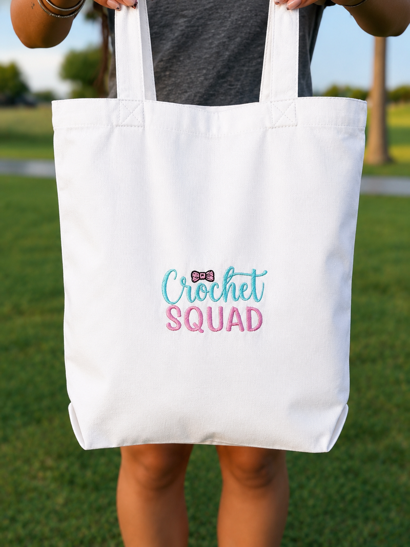 A white canvas tote bag with 'Crochet SQUAD' printed on the front in teal and pink lettering, held by a person outdoors on a grassy lawn.