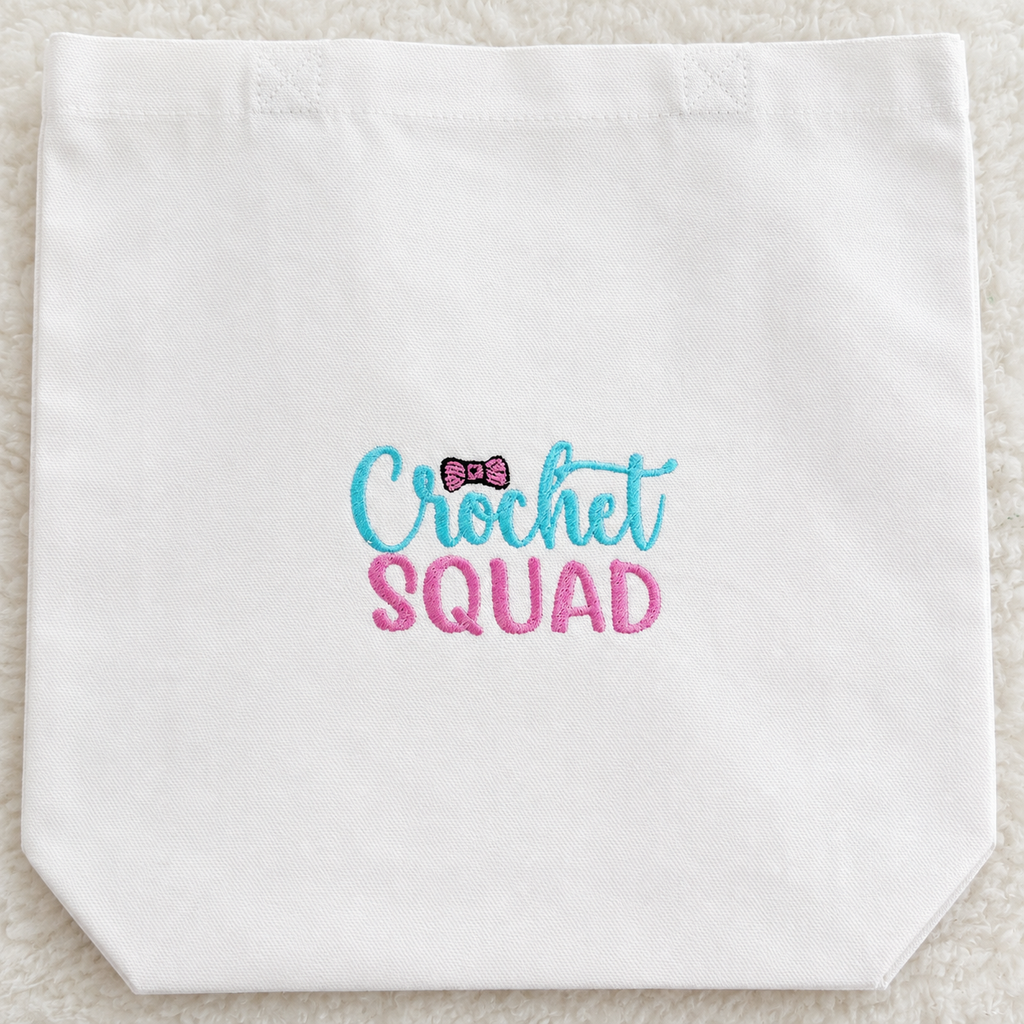 A white canvas tote bag with 'Crochet SQUAD' embroidered on the front in light blue and pink, featuring a small pink bow accent, laid flat on a white textured background.