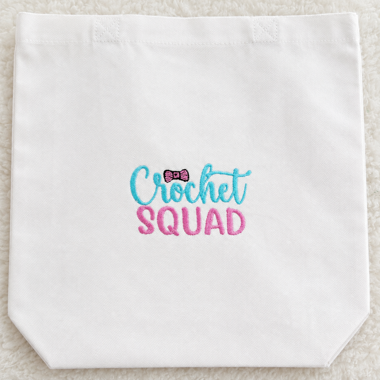 A white canvas tote bag with 'Crochet SQUAD' embroidered on the front in light blue and pink, featuring a small pink bow accent, laid flat on a white textured background.