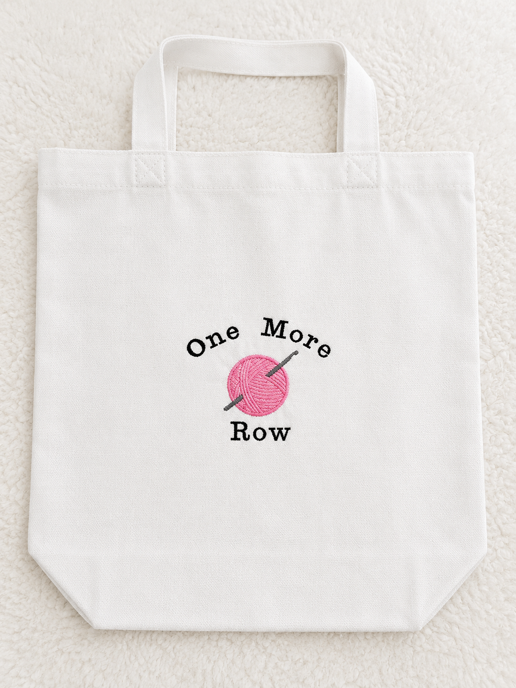 A white canvas tote bag with a printed graphic on the front that reads 'One More Row' around a pink ball of yarn with knitting needles, placed on a textured white fabric background.