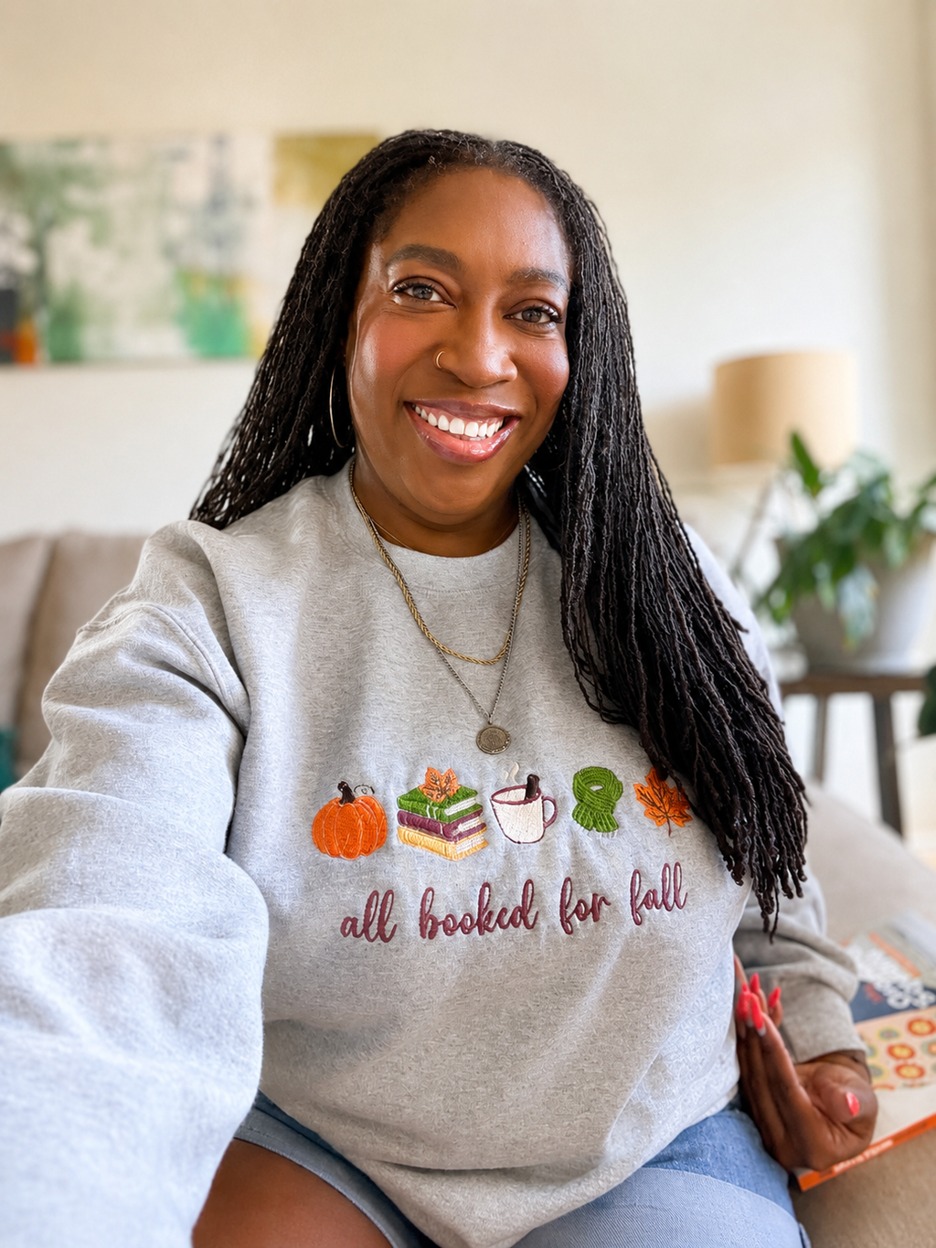 A smiling woman with long black locs sits indoors wearing a light heather gray sweatshirt with a fall-themed graphic that reads 'all booked for fall' with pumpkins, books, a mug, and a leaf. The background shows a softly blurred living room with a couch, lamp, and plant.
