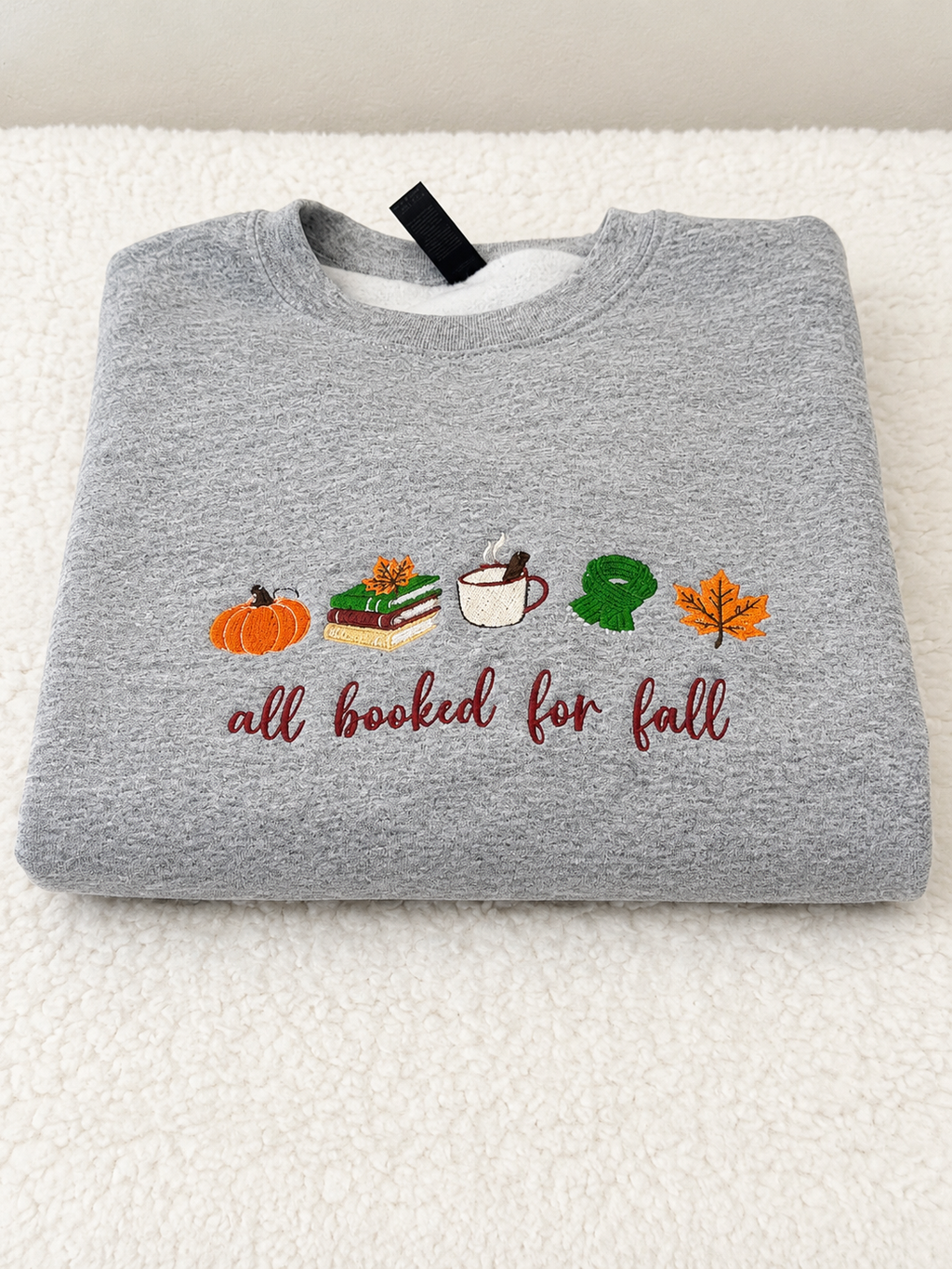 A folded heather gray sweatshirt on a white textured background, featuring an autumn-themed graphic with a pumpkin, books, coffee cup, green monster, maple leaf, and the text 'all booked for fall' in burgundy script.
