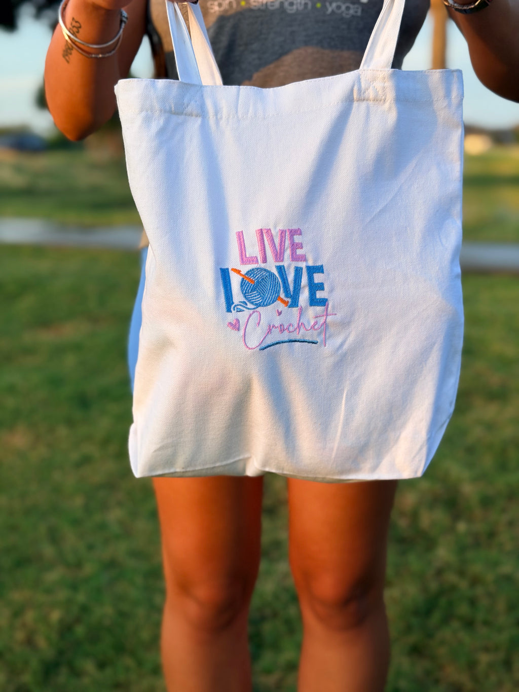 A white canvas tote bag with 'LIVE LOVE crochet' printed on the front in pink, blue, and purple, held by a person outdoors on a grassy lawn.