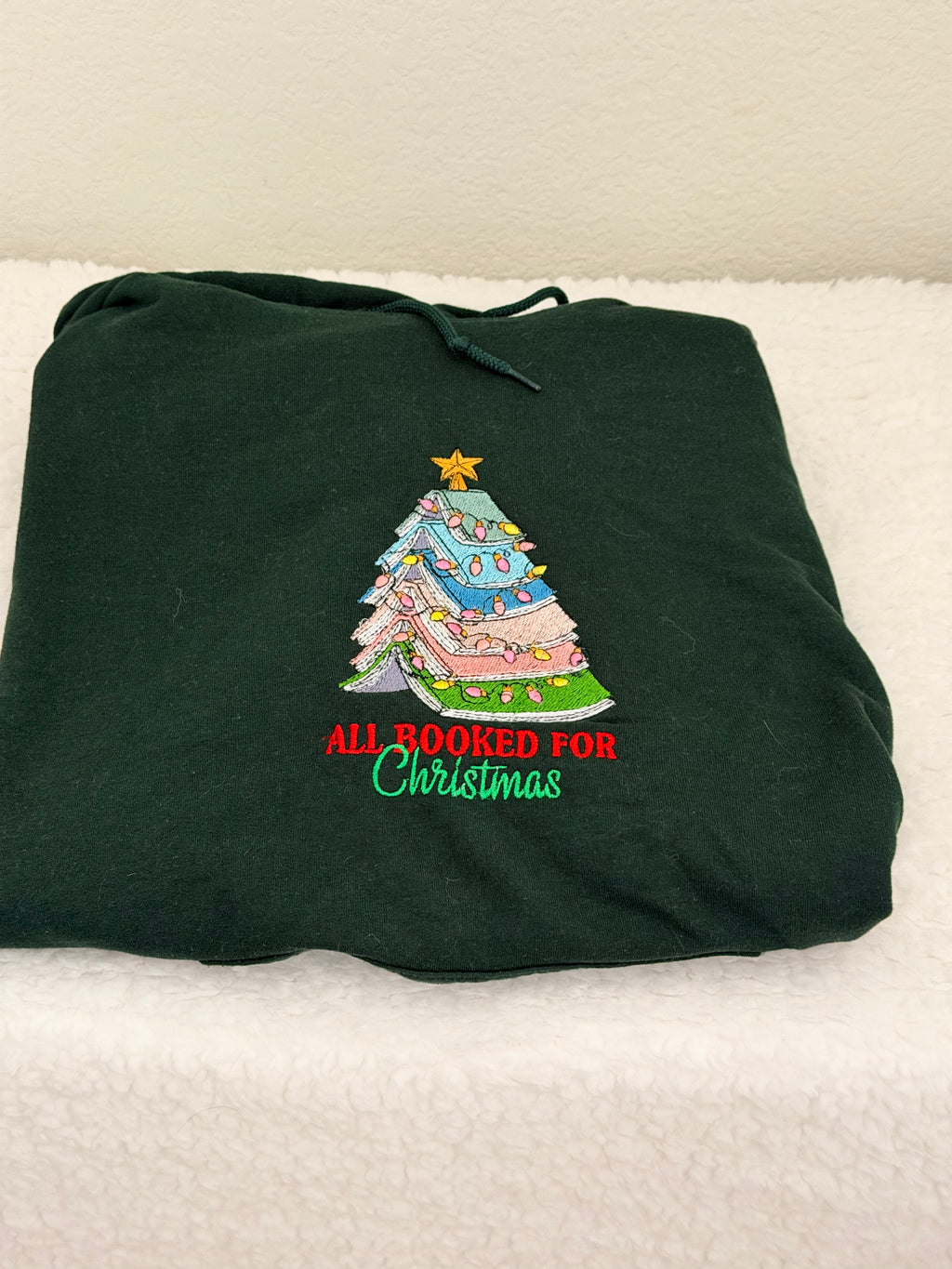 A dark forest green drawstring hoodie folded on a white textured surface, featuring a colorful embroidered Christmas tree and the text 'ALL BOOKED FOR Christmas' in red and green.