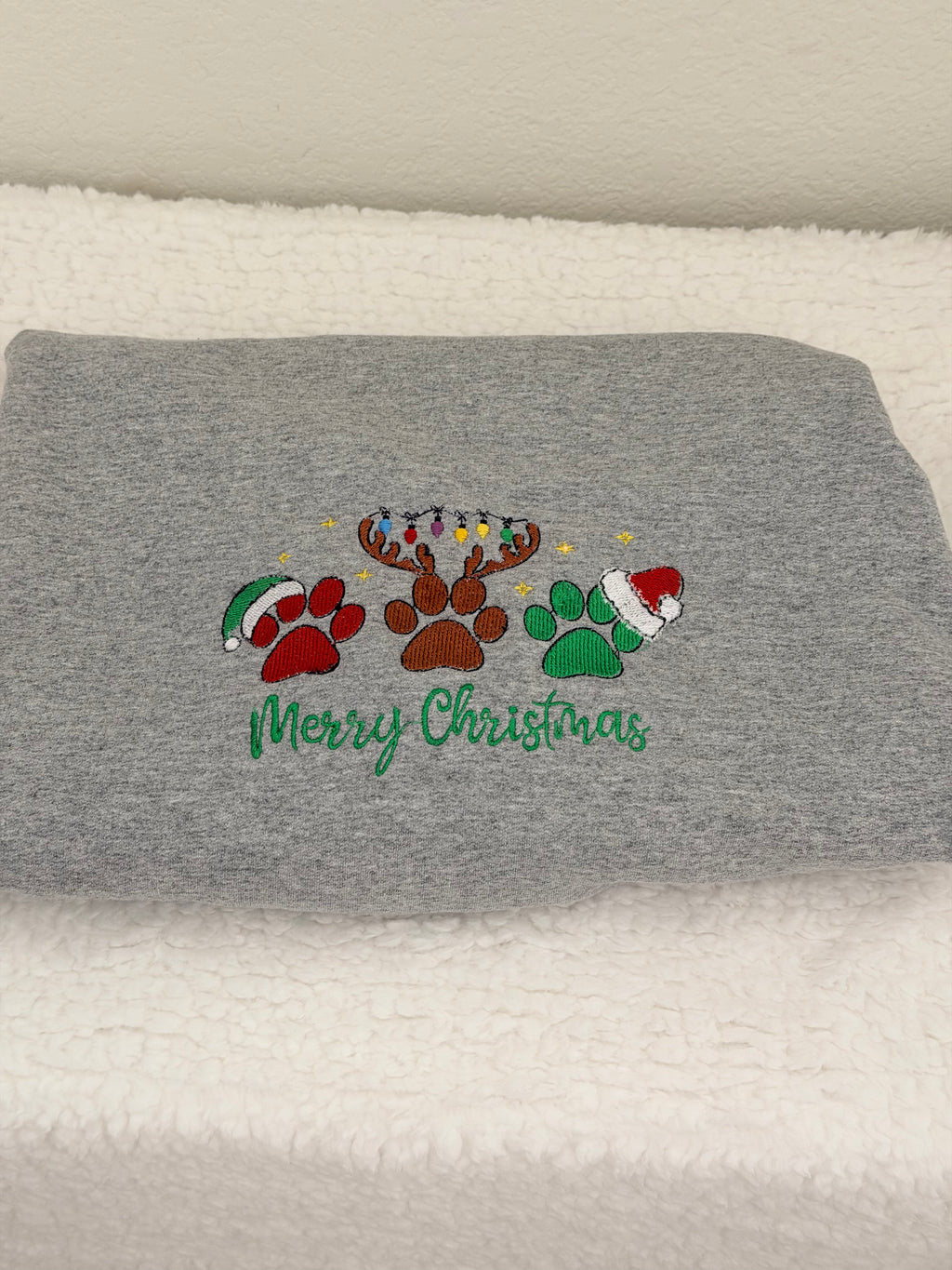A folded gray heather sweatshirt or hoodie resting on a white textured blanket. The front features a colorful embroidered design with a reindeer, paw prints, Christmas trees, and Santa hats above the green cursive text 'Merry Christmas'.