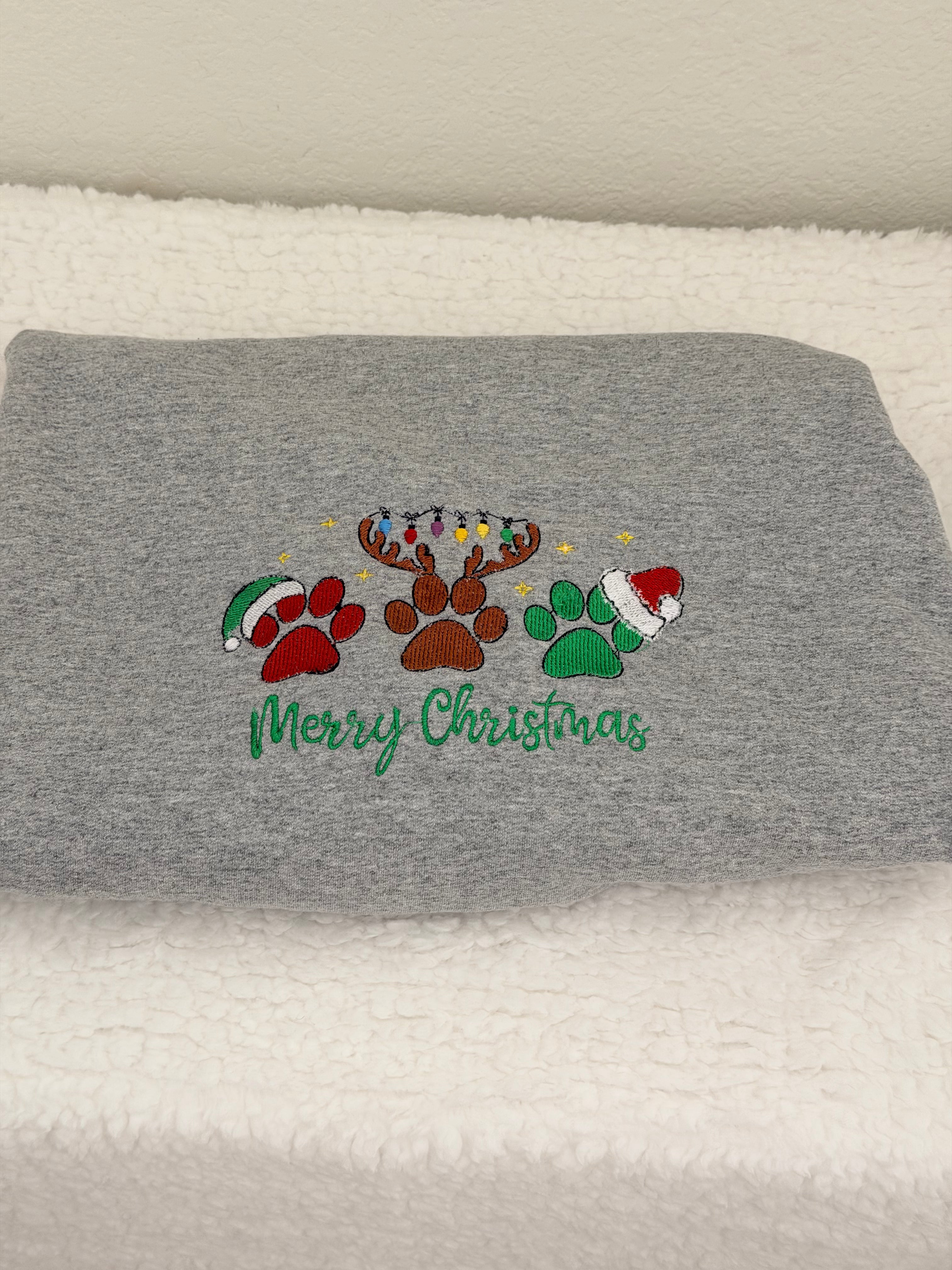 A folded gray heather sweatshirt or hoodie resting on a white textured blanket. The front features a colorful embroidered design with a reindeer, paw prints, Christmas trees, and Santa hats above the green cursive text 'Merry Christmas'.