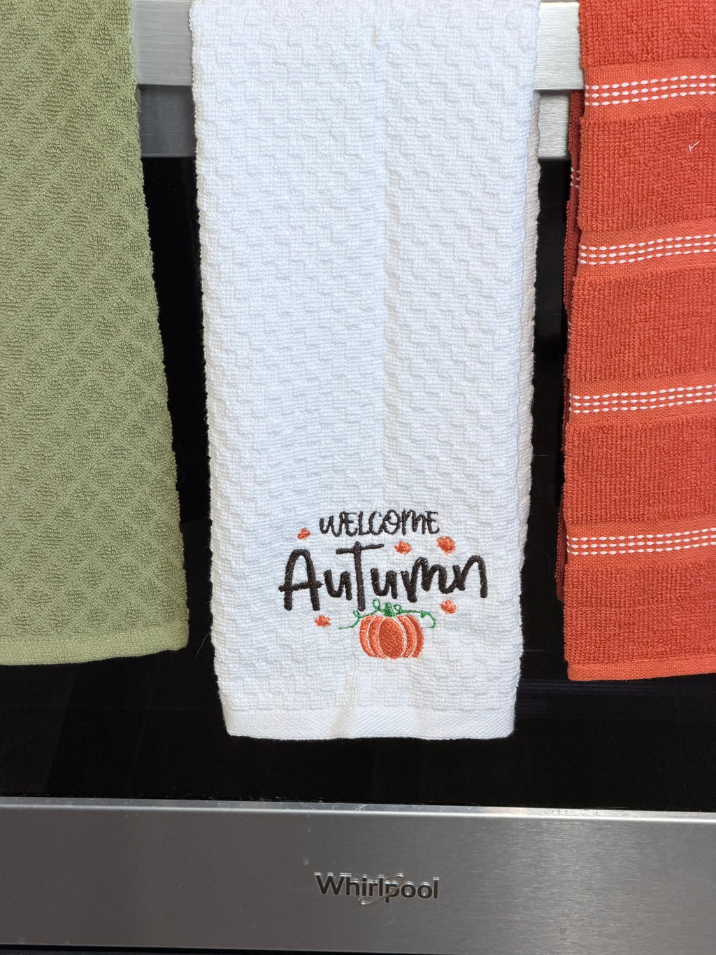 A white waffle-weave kitchen towel hanging on a rack, embroidered with 'Welcome Autumn' and a small pumpkin. Partially visible towels in green and orange hang beside it, and a stainless steel Whirlpool appliance is visible below.