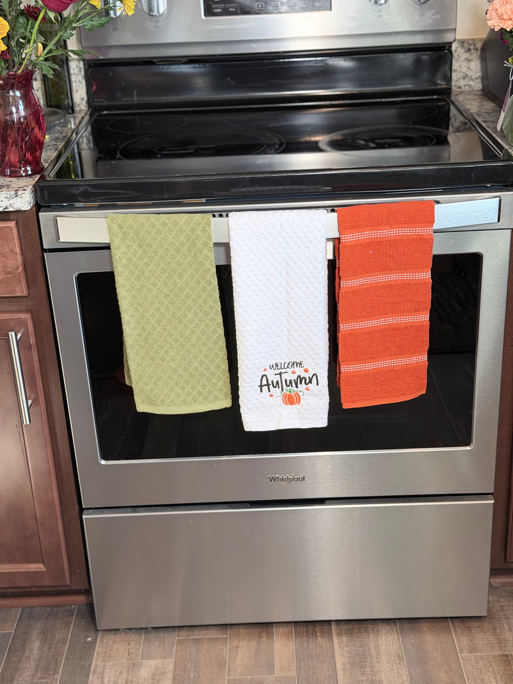 Three kitchen towels hanging on the handle of a stainless steel Whirlpool oven in a kitchen setting. The towels are sage green, white with 'Welcome Autumn' text and pumpkin graphic, and orange with thin white stripes.