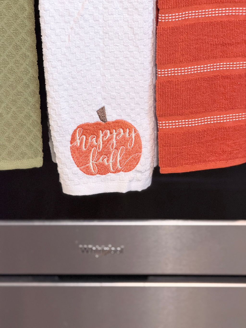 A white textured kitchen towel with an orange pumpkin graphic and the words 'happy fall' in white script, hanging next to other towels in green and orange in front of a stainless steel appliance.