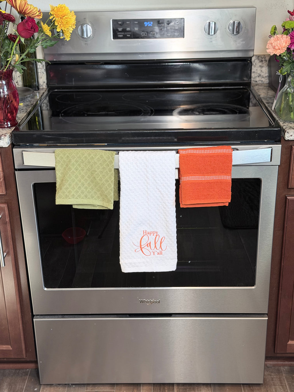 A stainless steel Whirlpool electric range with three kitchen towels hanging from the front handlebar in a kitchen setting. The center towel is white with 'Happy Fall' embroidered in orange, flanked by a green and orange towel.