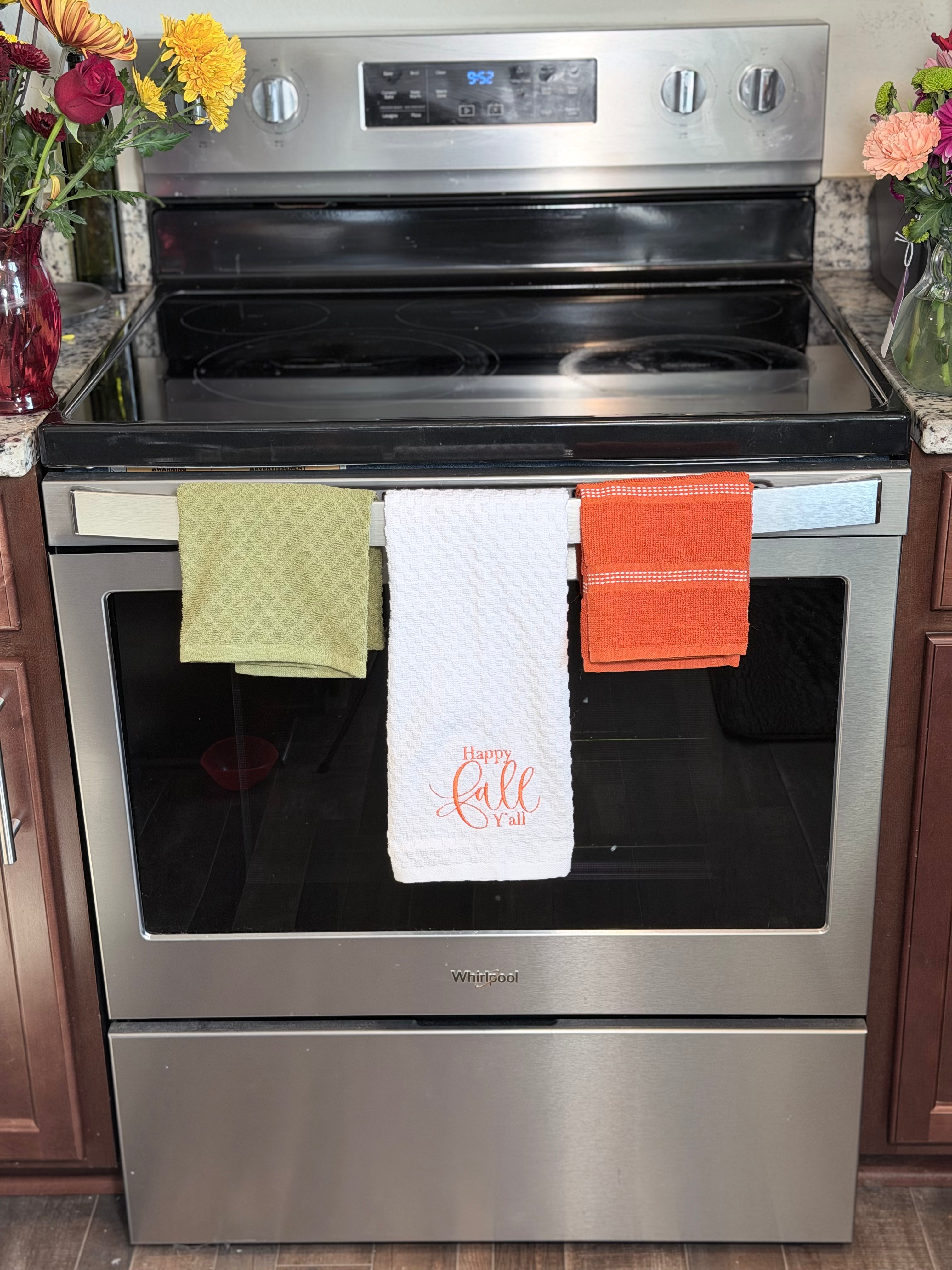 A stainless steel Whirlpool electric range with three kitchen towels hanging from the front handlebar in a kitchen setting. The center towel is white with 'Happy Fall' embroidered in orange, flanked by a green and orange towel.