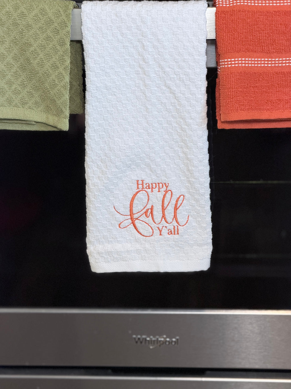 A white kitchen towel with 'Happy fall y'all' embroidered in red on the front, hanging on a rack in front of a black oven door. Other towels in green and orange are partially visible on either side.