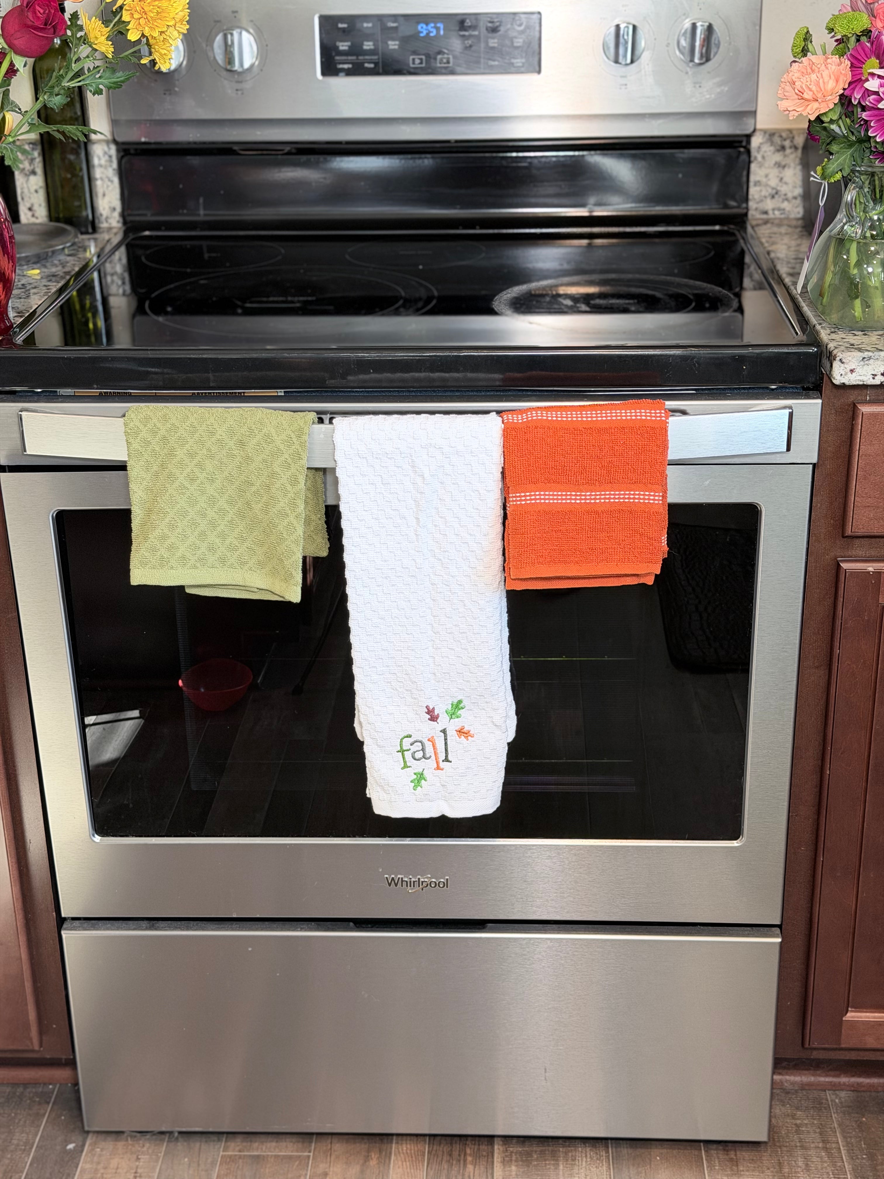 A stainless steel Whirlpool electric range with three kitchen towels hanging from the handle: a light green textured towel, a white towel with 'fall' embroidered in green and red, and an orange striped towel. The stove is set in a kitchen with wooden cabinets and flowers on the counters.