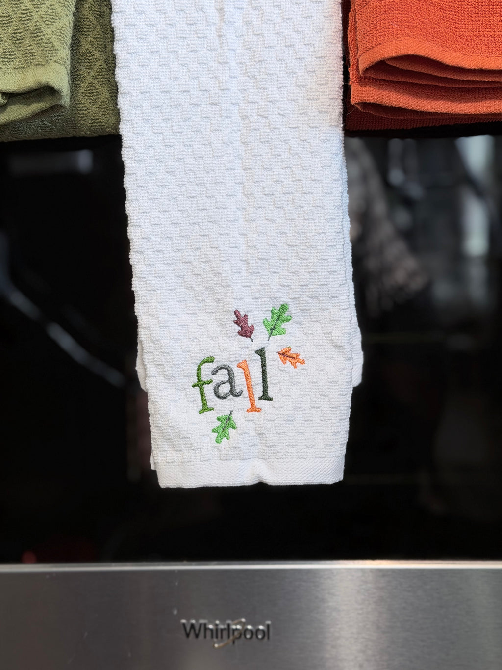A white waffle-weave kitchen towel hanging from a rack, embroidered with the word 'fall' in green and orange thread with small leaf accents. A Whirlpool appliance is visible below, and other towels in green and orange hang above.
