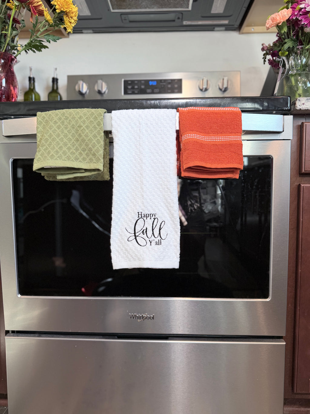 Three kitchen towels hanging on the handle of a stainless steel Whirlpool oven in a kitchen setting. The center towel is white with 'Happy Fall Furl' in black script, flanked by an olive green patterned towel on the left and an orange towel on the right.