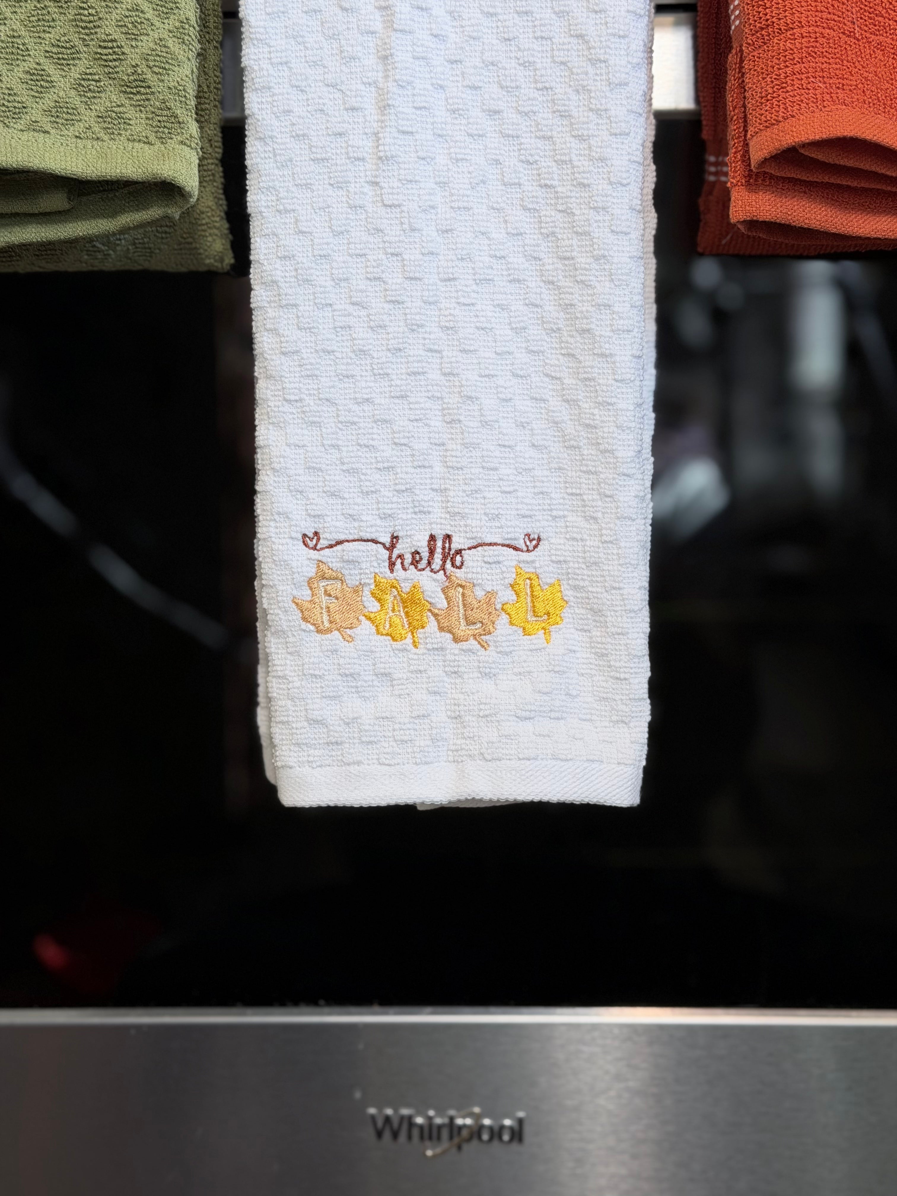 A white textured kitchen towel hanging in front of a black appliance, embroidered with the word 'hello' and autumn leaves in yellow, orange, and brown. A Whirlpool logo is visible on the appliance below.