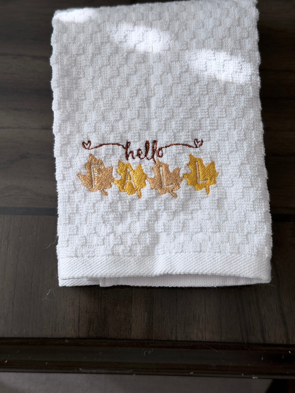 A folded white textured towel with 'hello' embroidered in brown above five autumn leaves in yellow, orange, and brown, resting on a dark wooden surface.