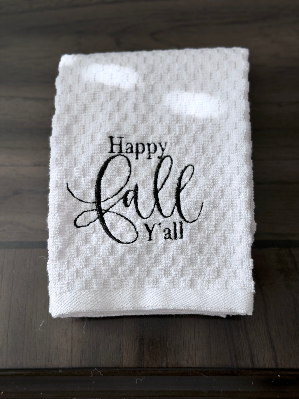 A folded white waffle-weave kitchen towel with the text 'Happy Fall Y'all' printed in black script, displayed on a dark wood surface.