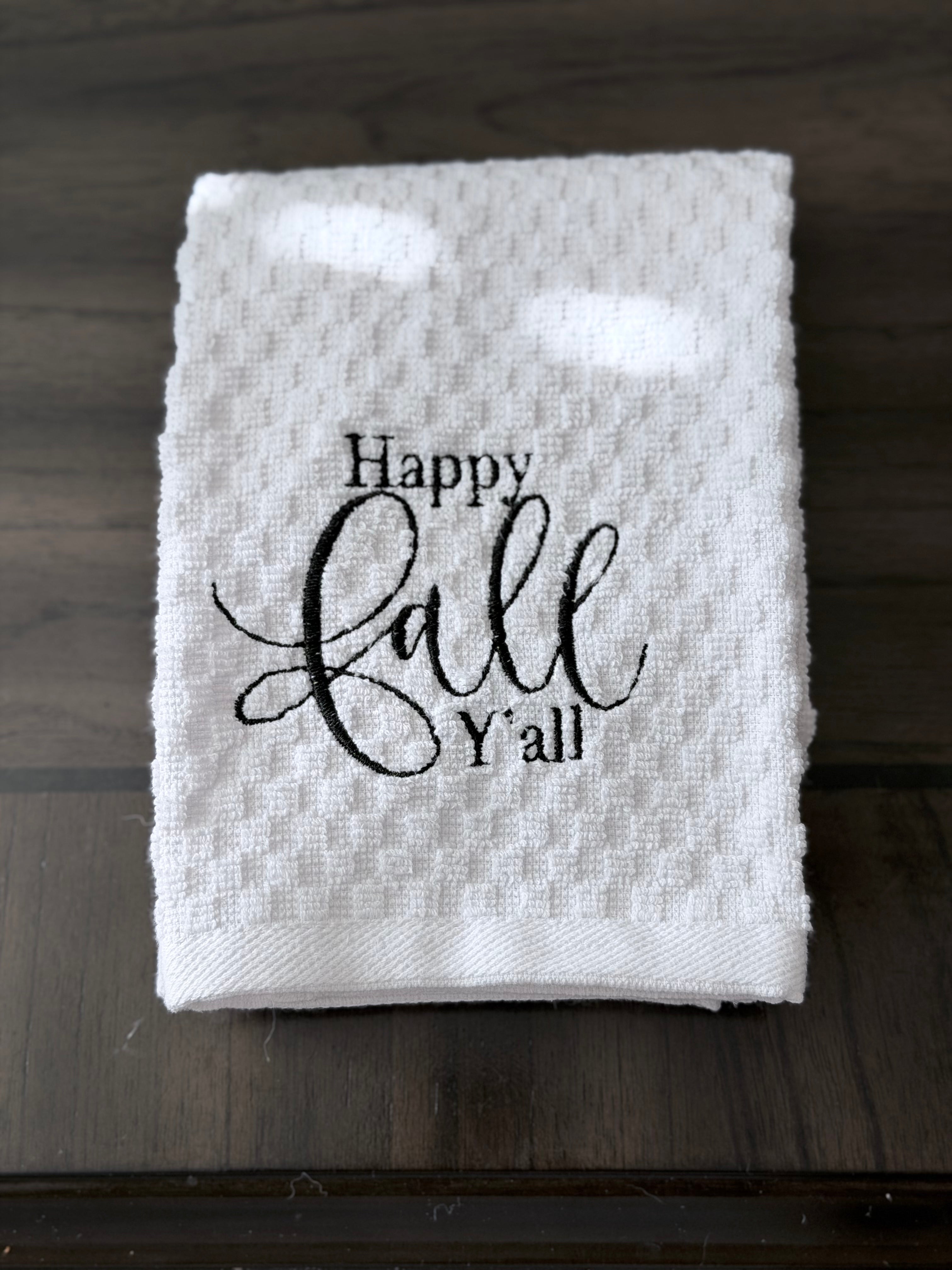 A folded white waffle-weave kitchen towel with the text 'Happy Fall Y'all' printed in black script, displayed on a dark wood surface.