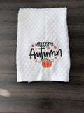 A white textured kitchen towel with 'WELCOME Autumn' embroidered in black, decorated with small orange leaves and a pumpkin, laid flat on a dark wood surface.