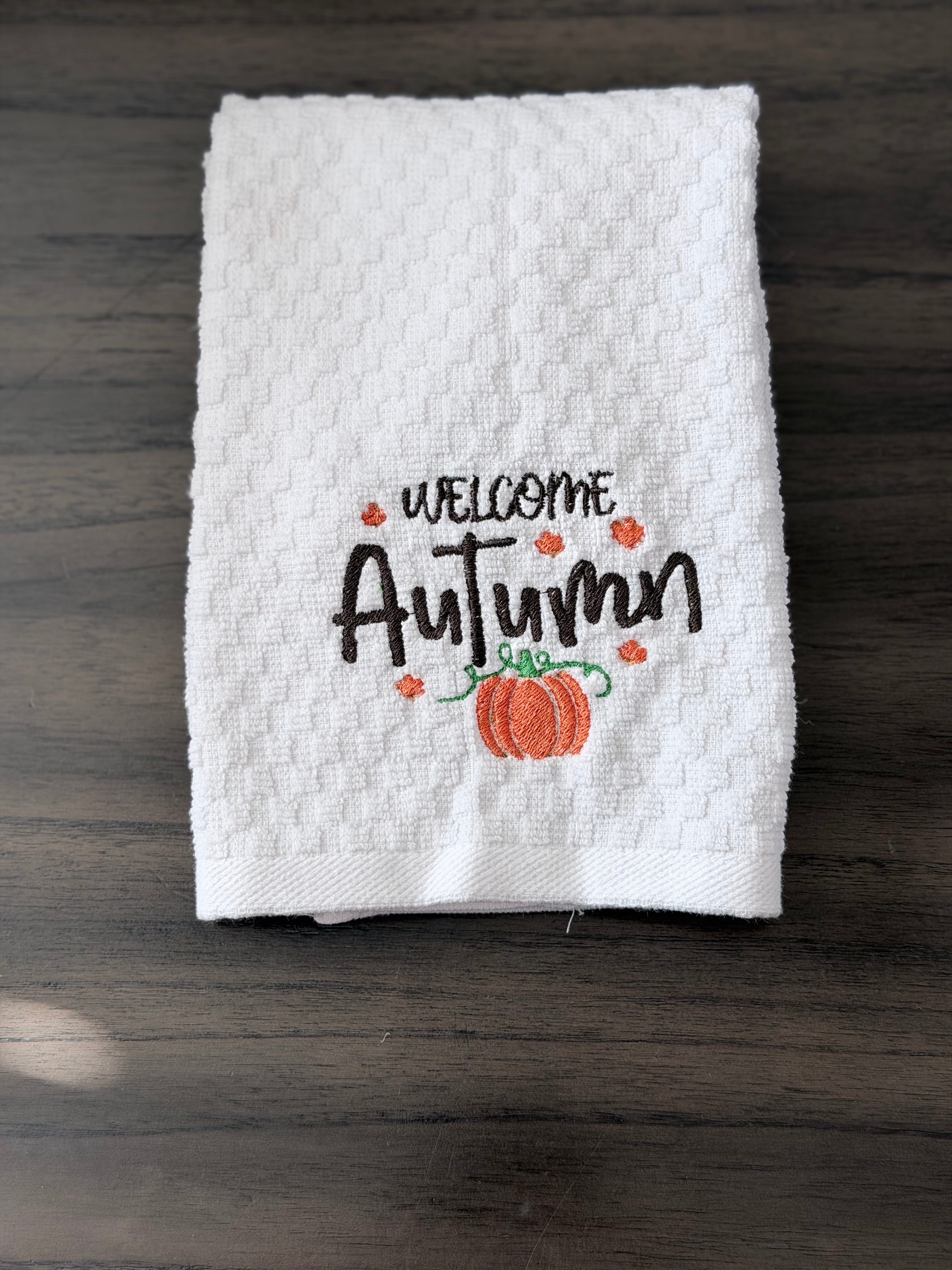 A white textured kitchen towel with 'WELCOME Autumn' embroidered in black, decorated with small orange leaves and a pumpkin, laid flat on a dark wood surface.