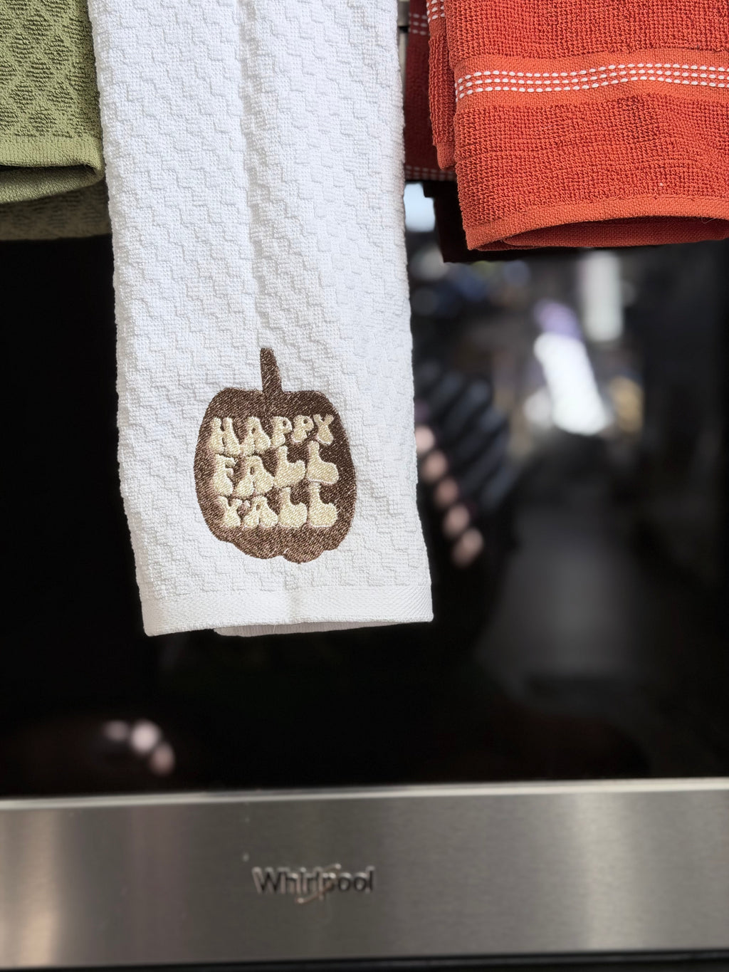 A white waffle-weave kitchen towel hanging in front of a stainless steel Whirlpool oven, featuring a brown embroidered pumpkin with the words 'Happy Fall Fall' in cream.
