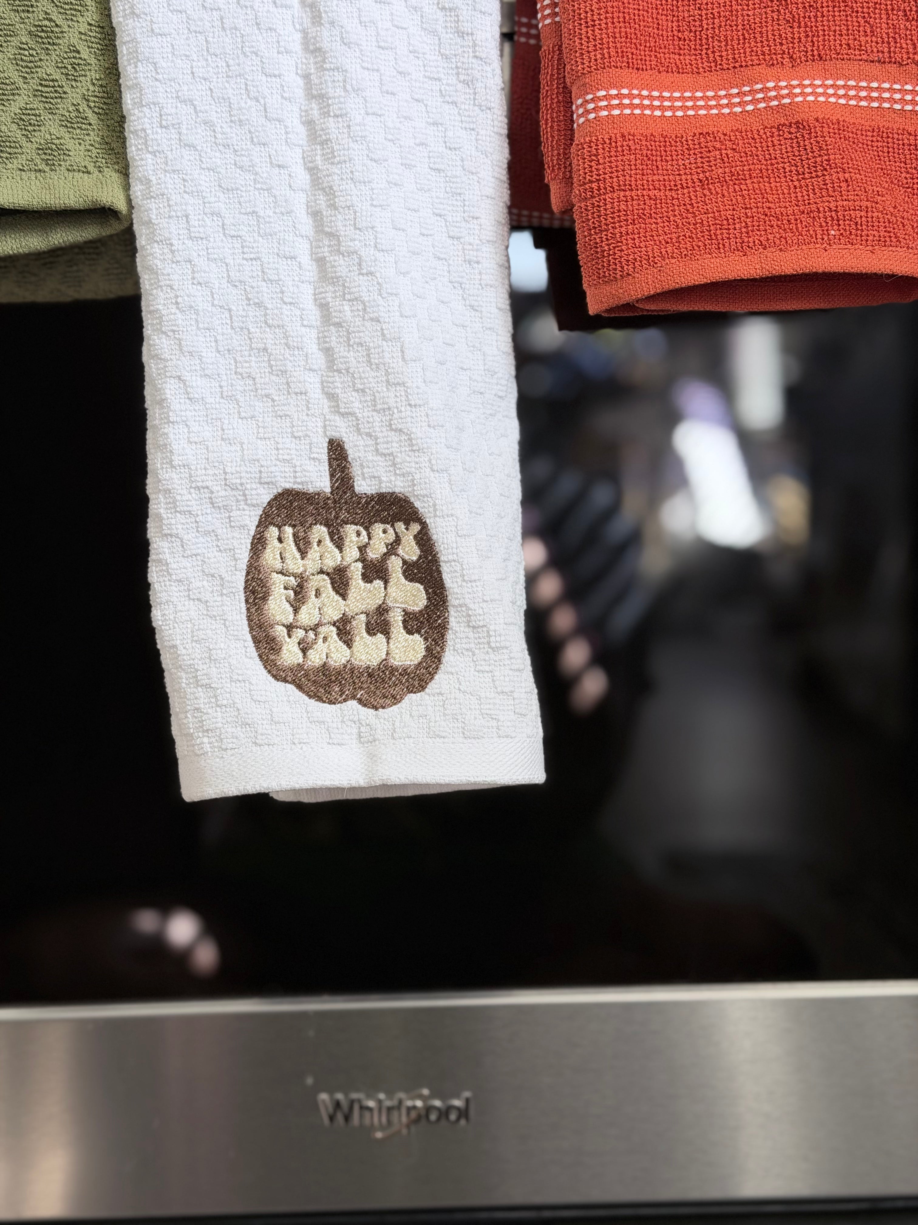 A white waffle-weave kitchen towel hanging in front of a stainless steel Whirlpool oven, featuring a brown embroidered pumpkin with the words 'Happy Fall Fall' in cream.
