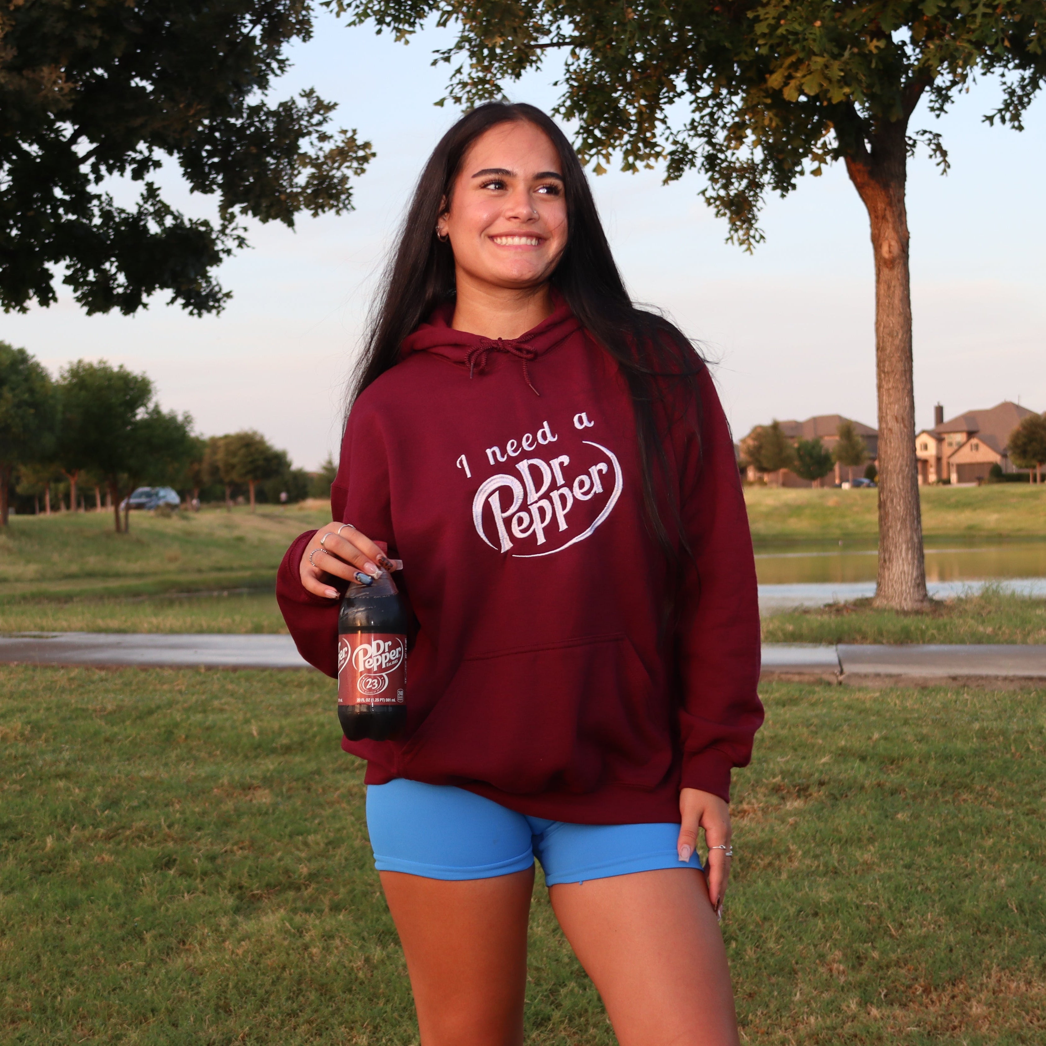 A woman standing outdoors in a grassy park area at sunset, wearing a maroon hoodie with a white 'I need a Pepper' graphic and blue shorts, holding a bottle of Dr Pepper.
