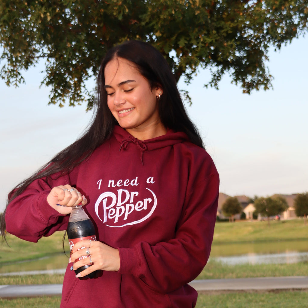 A woman outdoors on a grassy lawn wearing a maroon hoodie with the white text 'I need a Dr Pepper', holding a Dr Pepper bottle.