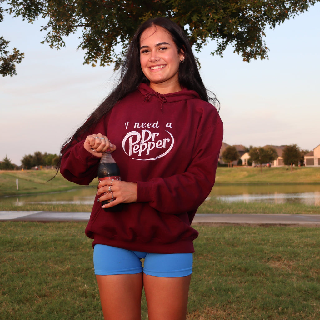 A woman standing outdoors in a grassy park area at sunset, wearing a maroon hoodie with a white 'I Need a Dr Pepper' graphic and blue shorts, holding a bottle.