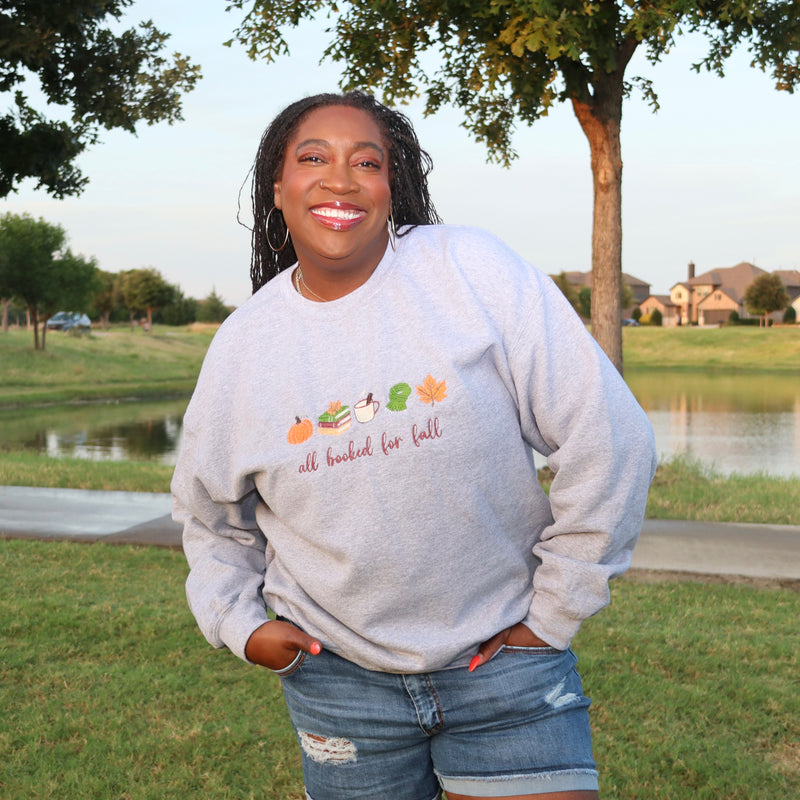 A smiling woman stands outdoors in a grassy park near a pond, wearing a light heather gray crewneck sweatshirt with a fall-themed graphic and the text 'all booked for fall' on the front, paired with denim shorts.
