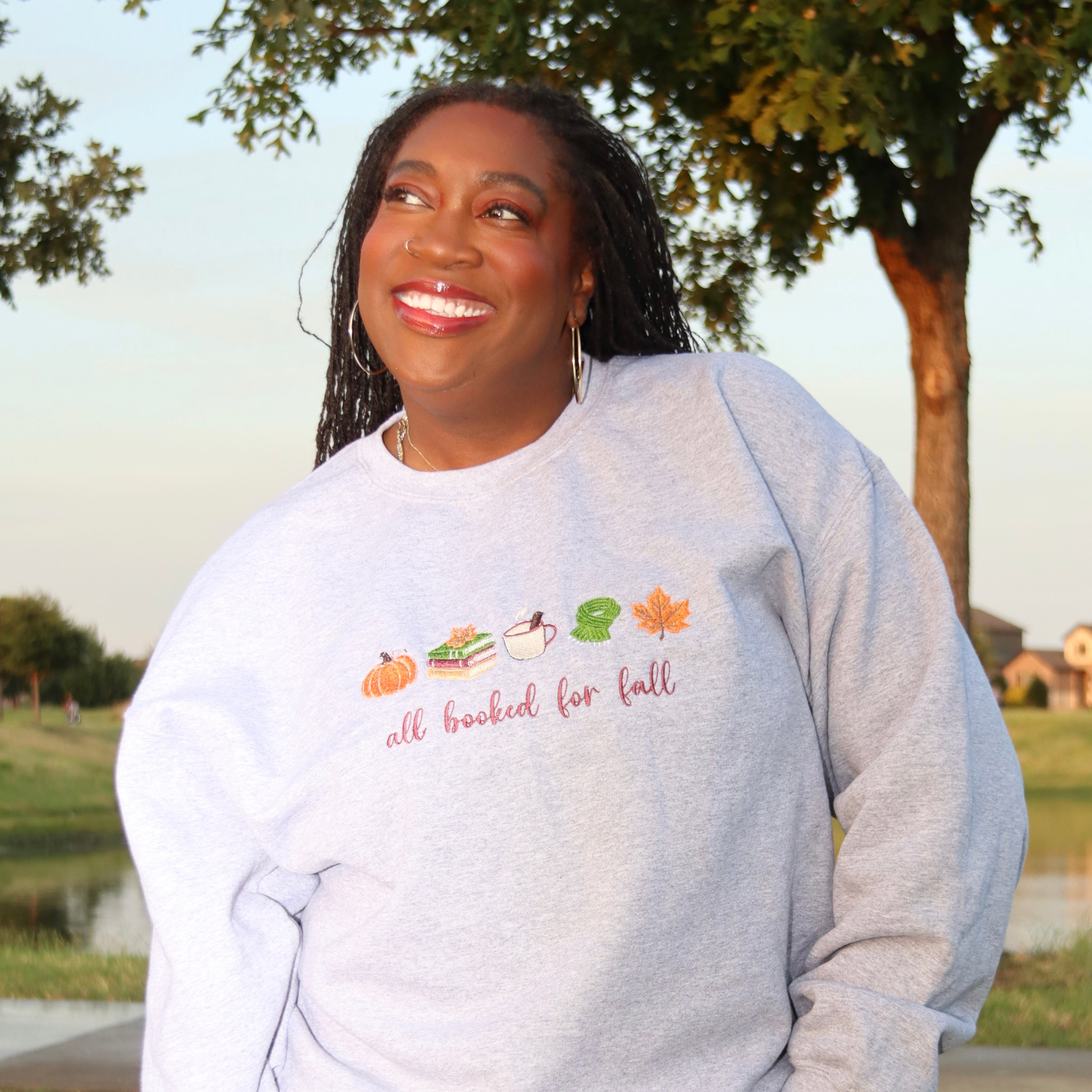 A smiling woman outdoors wearing a light heather gray sweatshirt with a colorful autumn-themed graphic and the text 'all booked for fall' across the chest.