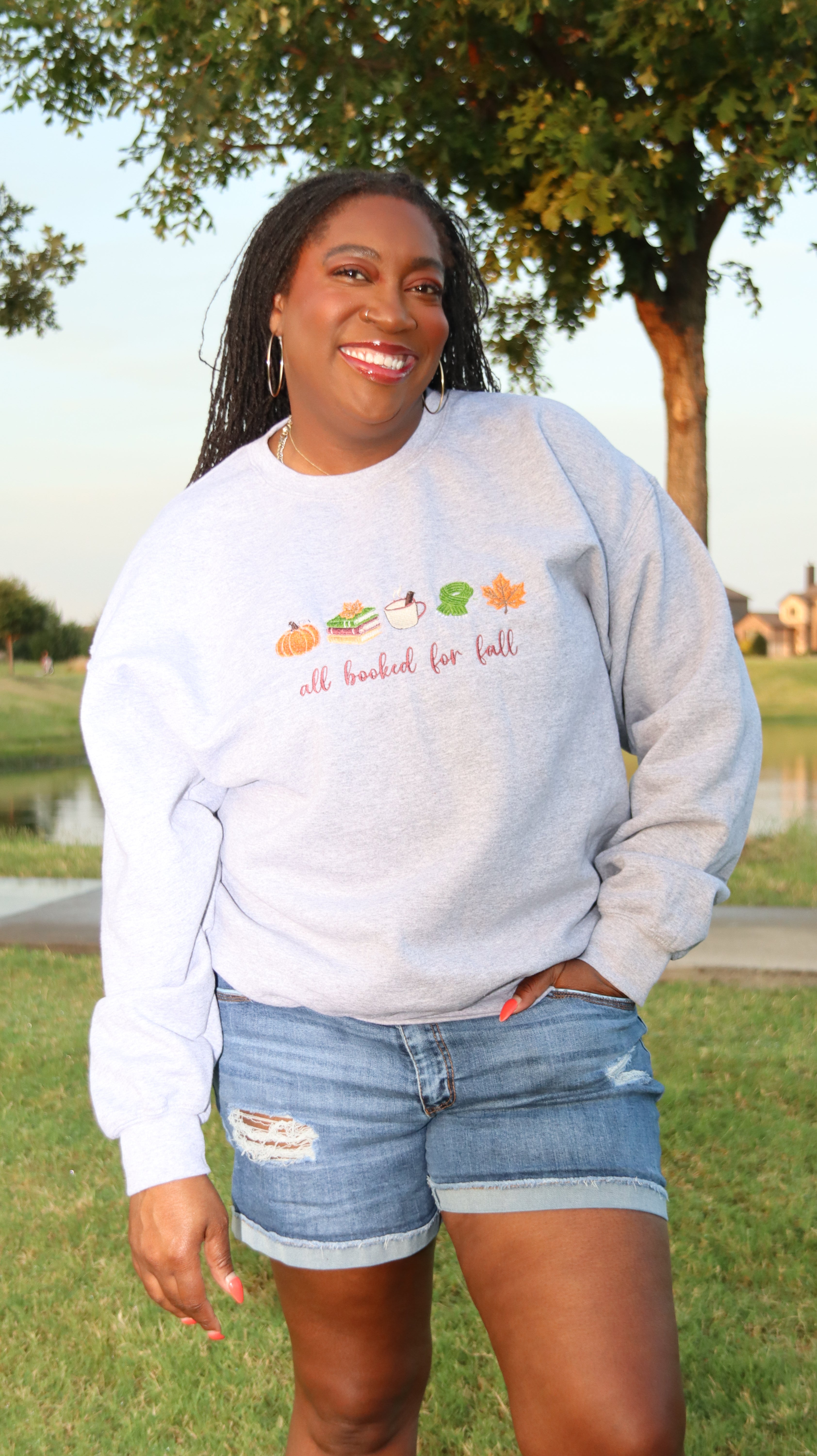A woman wearing a light heather gray sweatshirt with a fall-themed graphic and the text 'all looked for fall' paired with denim shorts, standing outdoors in a park setting.