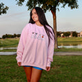 Smiling model wearing a light pink Ghost Dogs Trick-or-Treat embroidered crewneck sweatshirt featuring four adorable dogs dressed as ghosts, photographed outdoors during golden hour.