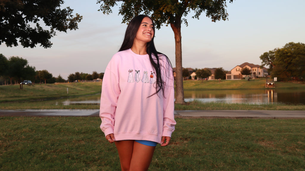 Model wearing a light pink Ghost Dogs Trick-or-Treat embroidered crewneck sweatshirt with four ghost dogs, standing outdoors near a lake at sunset.