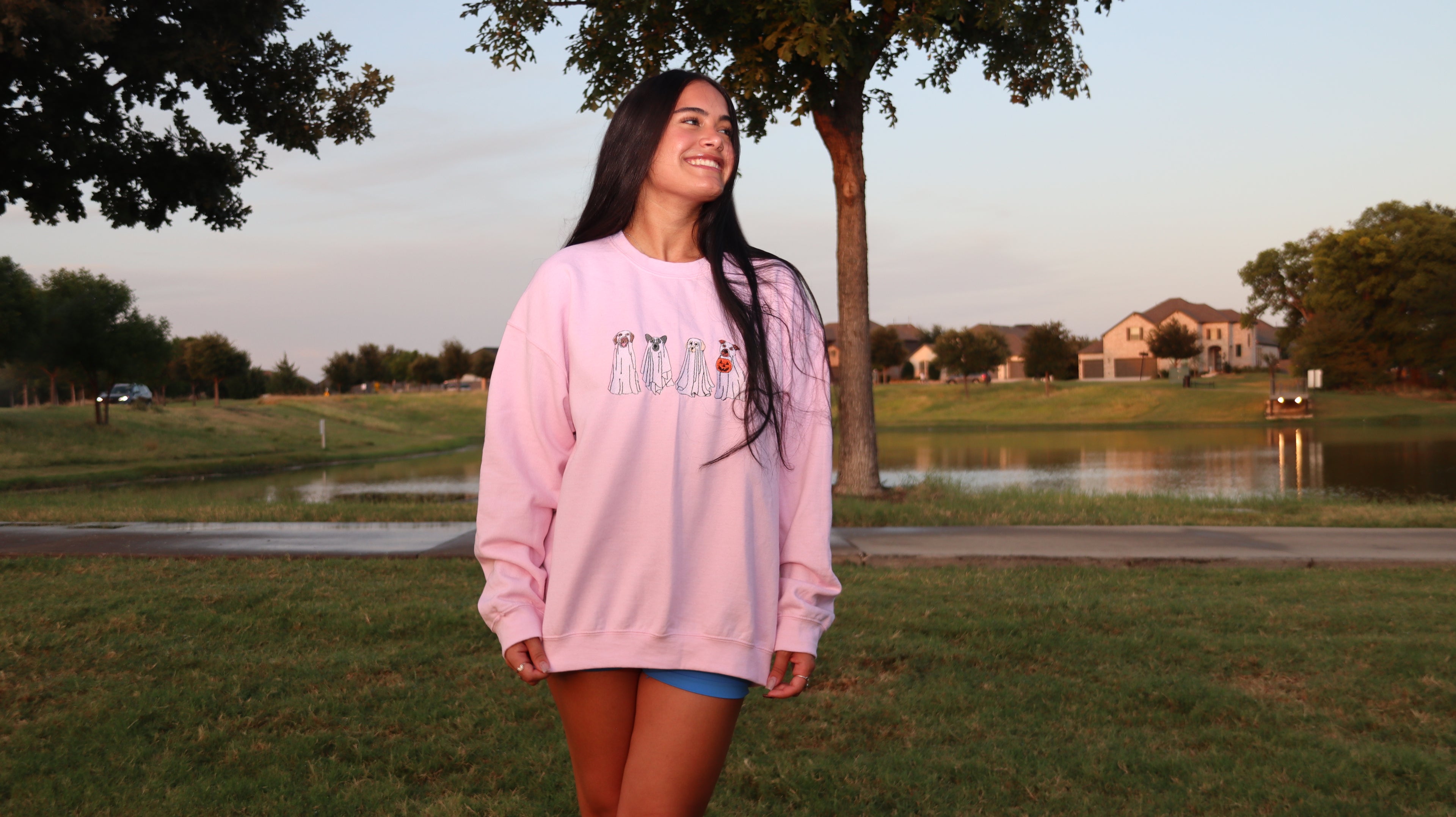 Model wearing a light pink Ghost Dogs Trick-or-Treat embroidered crewneck sweatshirt with four ghost dogs, standing outdoors near a lake at sunset.