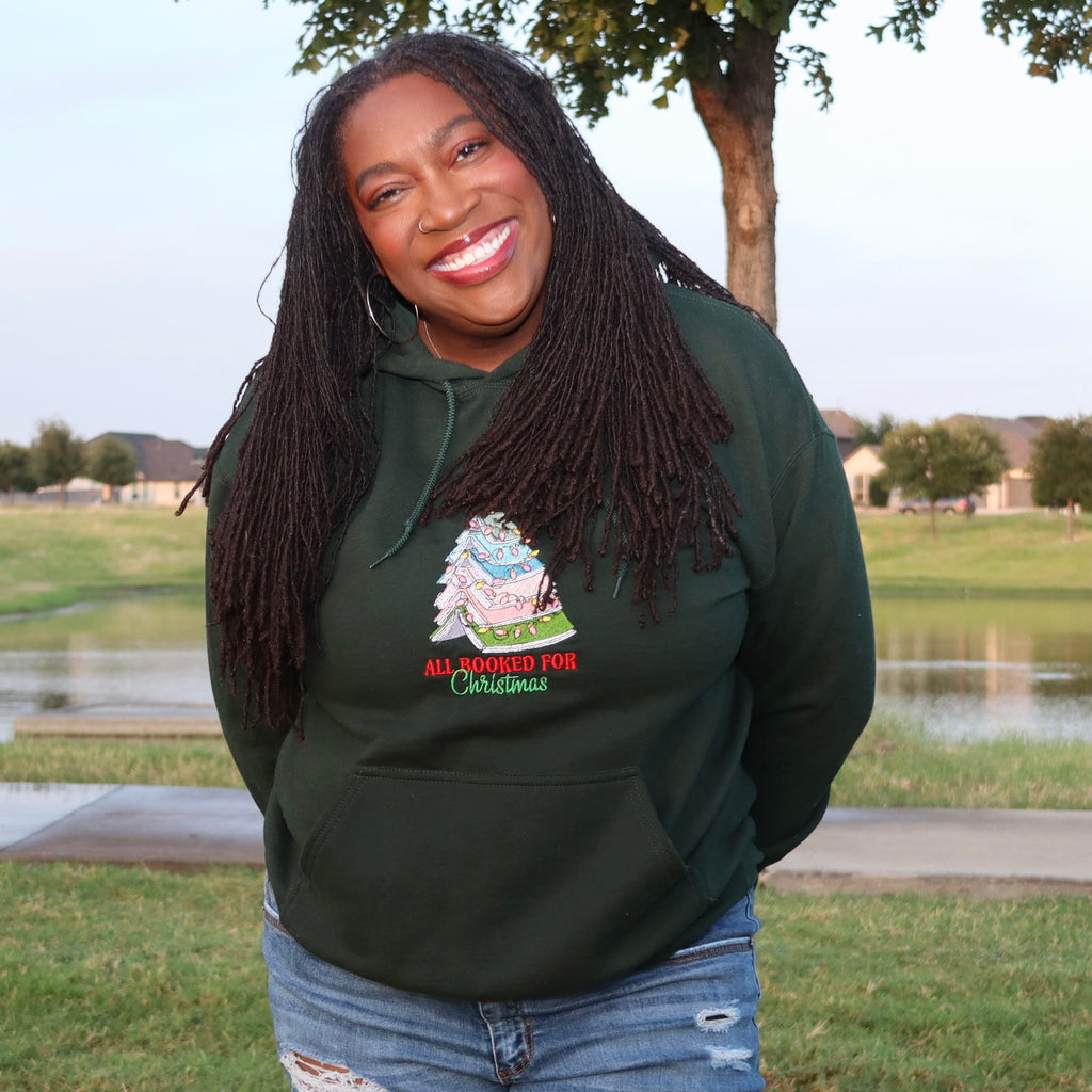 A smiling woman with long black dreadlocks stands outdoors wearing a dark green hoodie with a Christmas tree graphic and the text 'ALL-APPEARED FOR Christmas' on the front, paired with blue jeans. The background shows a grassy park area with a pond and trees.