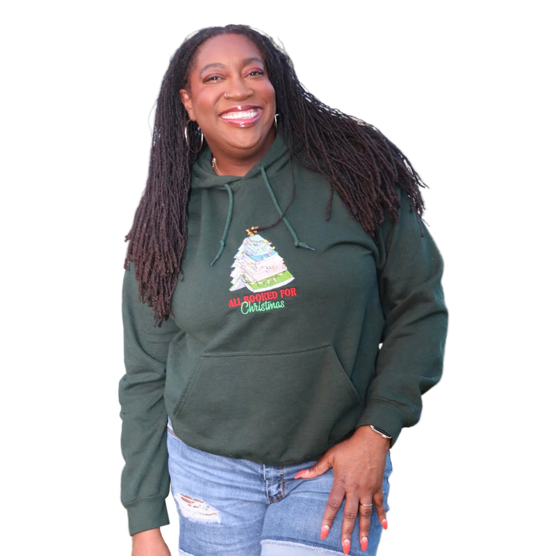 A smiling woman with long dark dreadlocks stands wearing a dark green pullover hoodie with a Christmas tree graphic and the words 'ALL MOURNED FOR Christmas' on the front, paired with light blue jeans. 