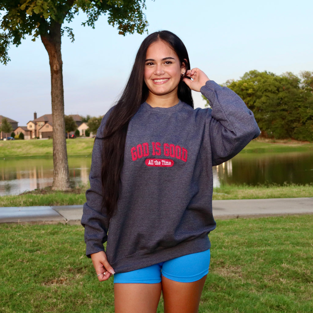 A woman standing outdoors in front of a lake and trees, wearing a dark heather gray sweatshirt with red text that reads 'GOD IS GOOD All Out There' and bright blue shorts.