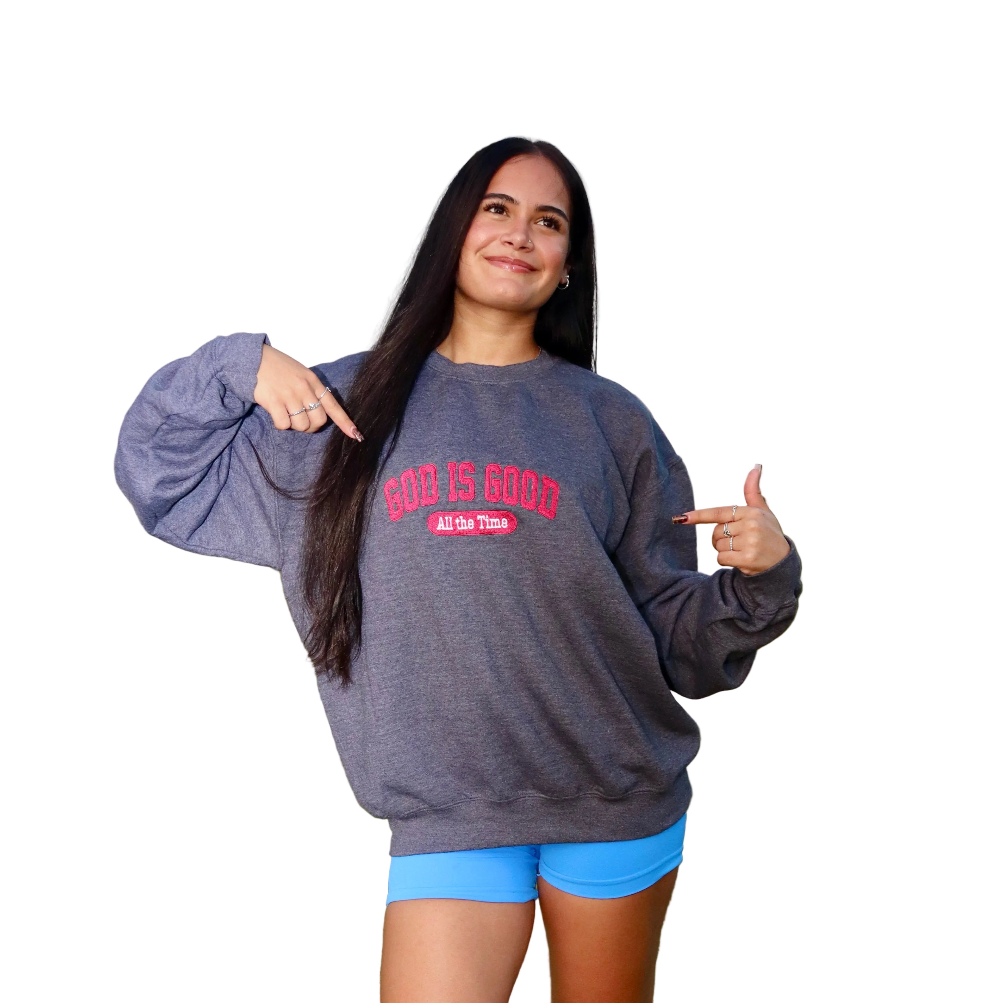 A woman standing wearing a dark heather gray sweatshirt with red text that reads 'GOD IS GOOD ALL GO THOSE' and bright blue shorts.