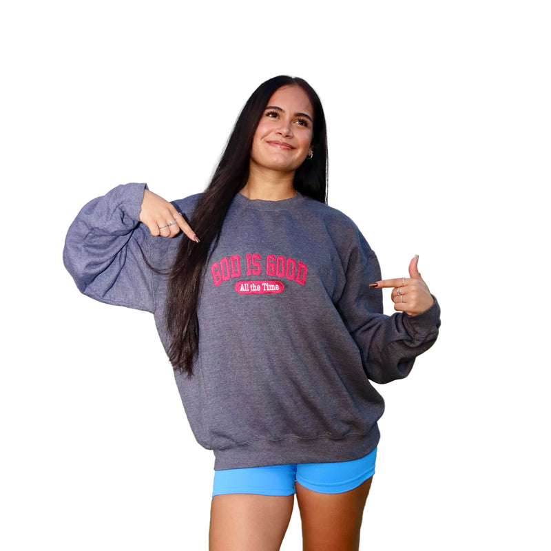 A woman standing wearing a dark heather gray sweatshirt with red text that reads 'GOD IS GOOD ALL GO THOSE' and bright blue shorts.