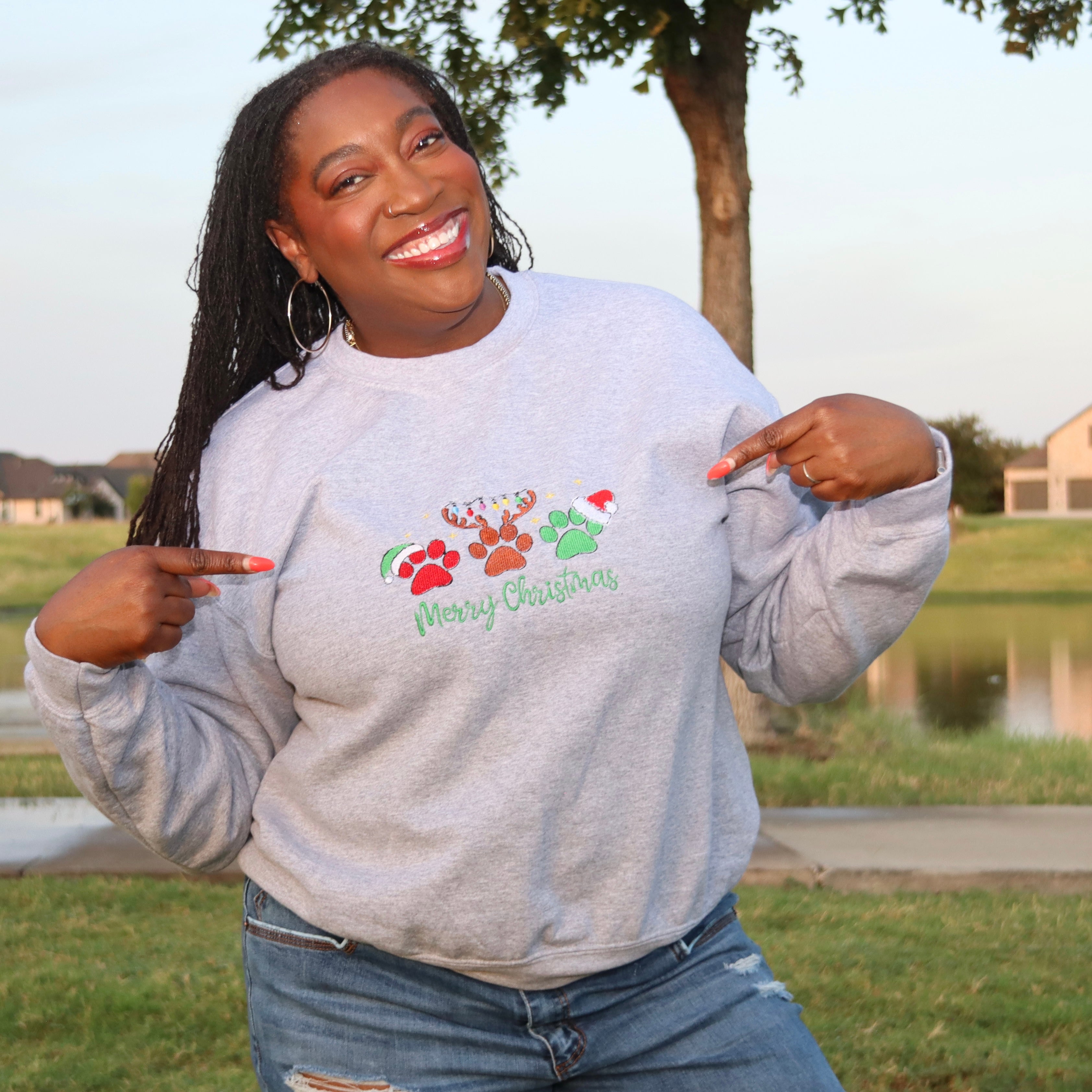 A woman wearing a light gray crewneck sweatshirt with a colorful Christmas graphic and the words 'Merry Christmas' printed on the front, posing outdoors in a grassy park area.
